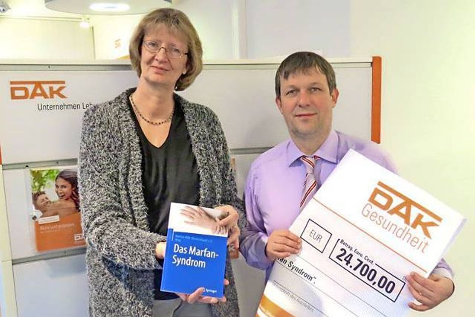 Nils Junker, Leiter des Servicezentrums der DAK, überreicht Marina Vogler (der Geschäftsstellenleiterin der Marfan Hilfe Deutschland e.V.) einen Scheck. Damit wird die Finanzierung des Buchprojekts „Das Marfan-Syndrom“ unterstützt.