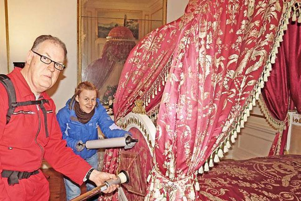 Vom Parade-Bett wirbelt Ulrike Unger mit einem Blasebalg den Staub auf, Restaurator Markus Freitag fängt  ihn dann mit dem Staubsauger auf, in dem sich ein Filter befindet, so dass der Staub nicht wieder mit der Luft nach draußen entweichen kann.