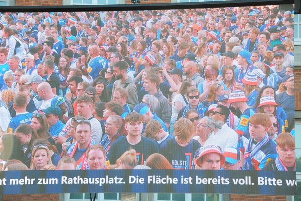 Auf einem der Großbildschirme ist gerade zu sehen, wie Fans dicht gedrängt auf die Ankunft der Mannschaft warten.