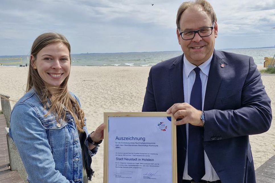 Bürgermeister Mirko Spieckermann und Nachhaltigkeitsmanagerin Lina Sophie Koop mit der Auszeichnung für die erfolgreiche Projektteilnahme „BNK bundesweit 2024“, in dessen Rahmen die Berichtserstellung nach dem Berichtsnahmen Nachhaltige Kommune (BNK) erfolgte. (Foto: Stadt Neustadt in Holstein)