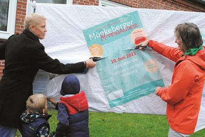 Jessica Salewski und Bürgermeisterin Hildegard Mersmann zerschneiden die Einweg-Tischdecken, mit denen der Schaukasten vor allzu neugierigen Blicken eingepackt war. Kinder haben sichtlich Spaß an der Enthüllung.