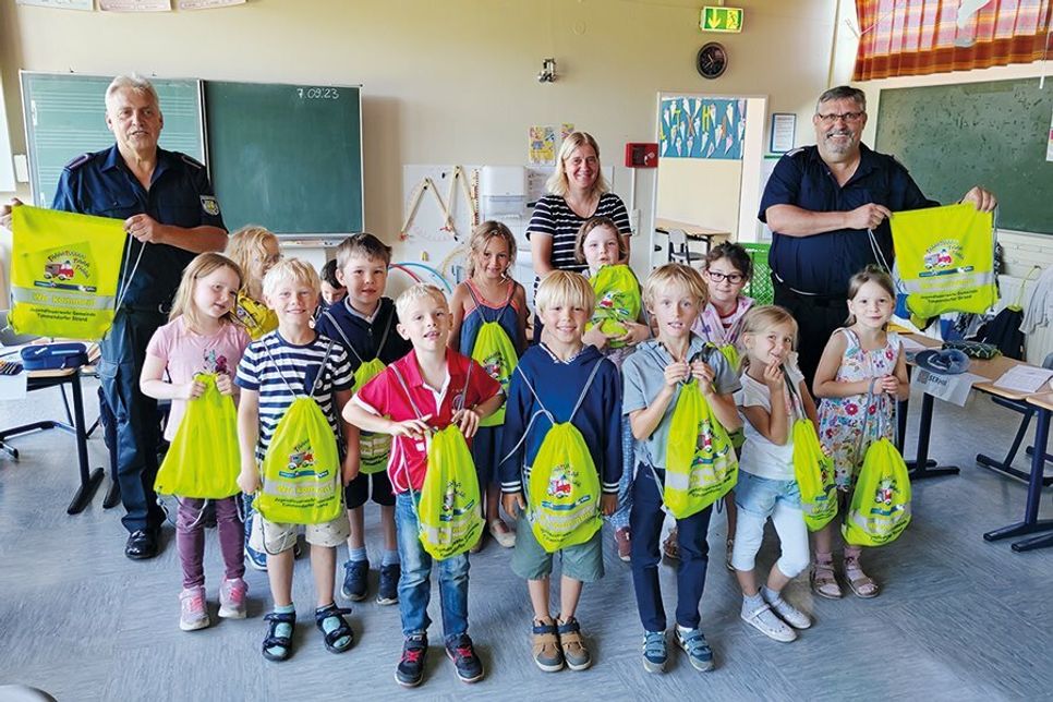 Stolz zeigen einige Timmendorfer Erstklässler der Klasse 1a mit ihrer Klassenlehrerin Veronika Krause ihre hell leuchtenden, gefüllten Turnbeutel, dahinter Thomas Scharbau (rechts) und Karl-Heinz Zellien von der Feuerwehr.