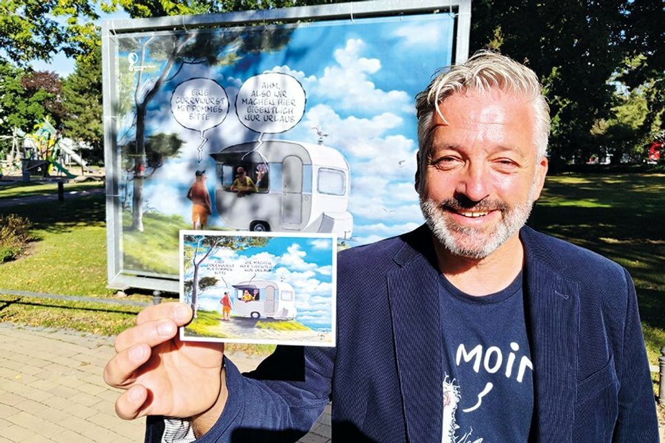 Cartoonist Frank Bahr mit einer zur Ausstellung gehörenden Postkarte vor einem seiner Werke.