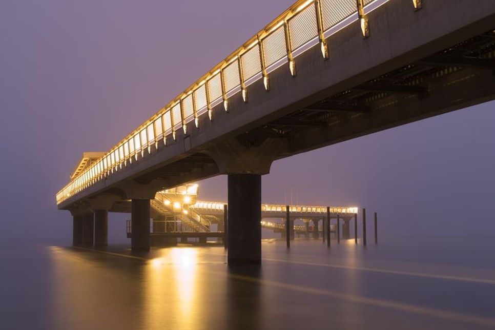 Anke Jeggle zeigt die Haffkruger Seebrücke in einer besonderen Atmosphäre. Die warmen Lichter spiegeln sich sanft im Wasser, während die Umgebung im Nebel versinkt.