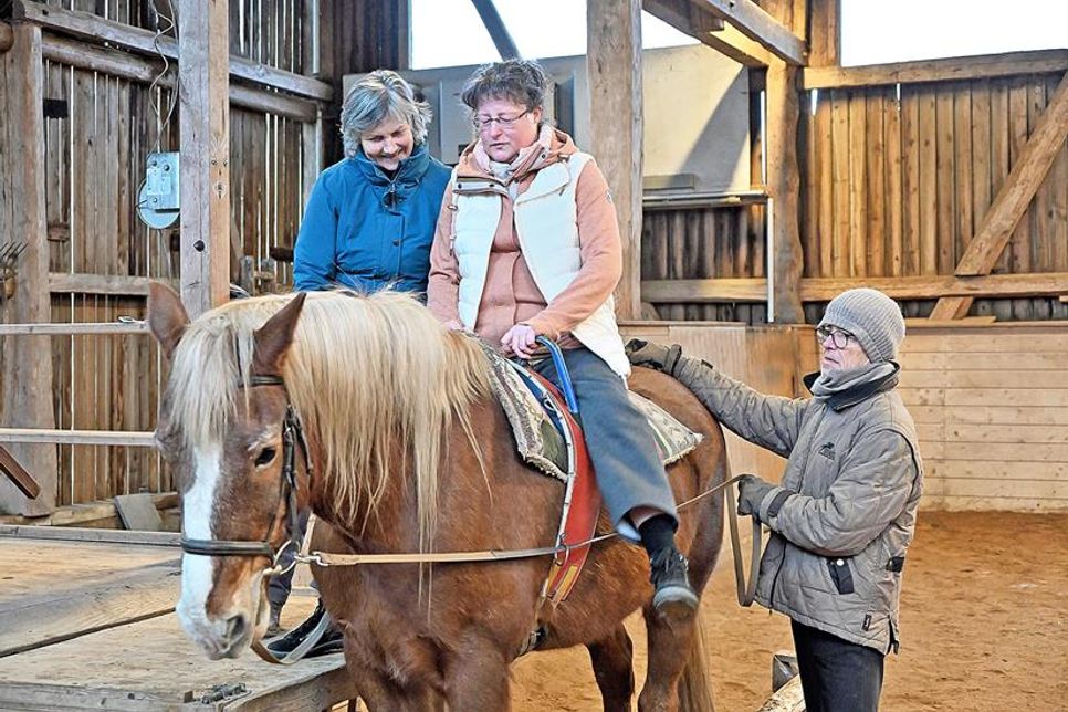 Auf Höhe des Pferdes kann sich Sabine Henning mit Unterstützung von Therapeutin Amelié von Bülow-Sartory vorsichtig auf dessen Rücken gleiten lassen. Das erfahrene Therapiepferd „Fidel“ an der Hand von Rüdiger Sartory bleibt ruhig stehen.