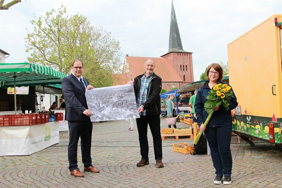 Bürgermeister Mirko Spieckermann (lks.) und Marktmeister Peter Heuer mit einem historischen Foto vom Wochenmarkt und Stadtmarketingkoordinatorin Andrea Brunhöber mit Blumen fürs – beziehungsweise vom – Geburtstagskind.