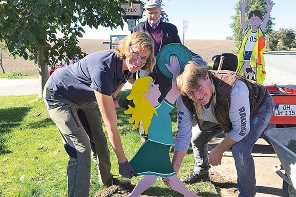 Ulrike Hansen und Torsten Köberich vom „Dorf-Team“ stellen eine der vier Holzfiguren auf. Im Hintergrund beobachtet Fiete Drückhammer das Geschehen.