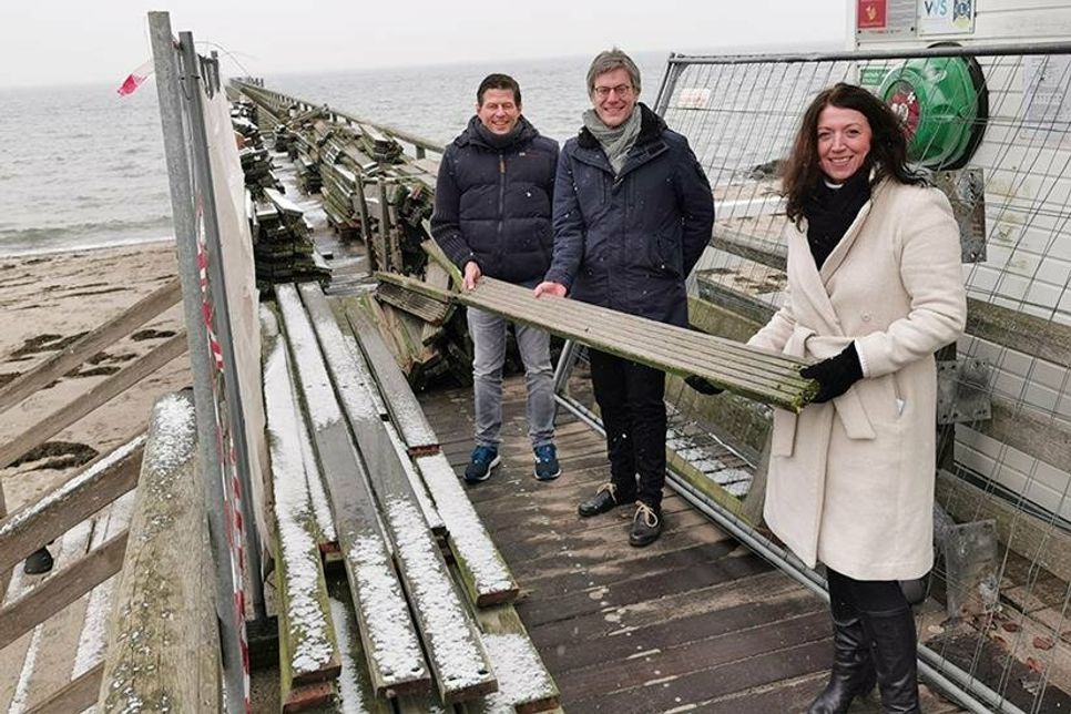 Thomas Albertin (Bauamt der Gemeinde Scharbeutz, links), TALB-Vorstand André Rosinski und Bürgermeisterin Bettina Schäfer mit einer bereits abmontierten Holzplanke, die man mit Glück als Erinnerungsstück erhalten kann.