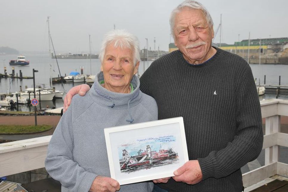 Rose-Marie und Bruno Schmidt auf der Terrasse ihres Wassersportclubs in Wellingdorf. Sie zeigen ein Bild, das typisch ist für Bruno Schmidt: Titel und Erläuterungen als Bestandteil eines Kunstwerks. Hier: „Der gesprengte U-Bootbunker, erste Sprengung 25. Oktober 1946“. Heute befindet sich an dieser Stelle der Ostuferhafen. Einen Teil davon kann man im Hintergrund der Aufnahme erkennen.