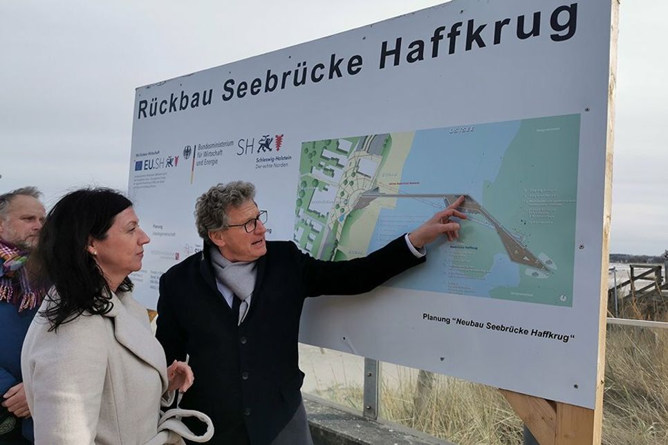 Bettina Schäfer, Bürgermeisterin der Gemeinde Scharbeutz, schaut sich mit Minister Dr. Bernd Buchholz die Bau-Tafel der neuen Seebrücke an.