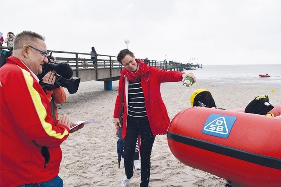 Verena Mertens tauft das neue DLRG-Rettungsboot mit Sekt auf den Namen ihres Vaters, beobachtet von DLRG-Spendensammler Achim Wiese und einem NDR-Fernsehteam.