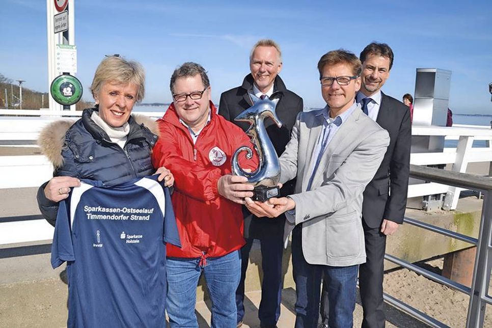 Sabine Bremeyer und Udo Brechtel vom NTSV präsentieren mit Tourismuschef Joachim Nitz das Laufshirt und einen der begehrten Seepferdchen-Pokale, sehr zur Freude von Fred Bobsin (hi.li.) und Volker Schinkewitz von der Sparkasse Holstein. (Foto: René Kleinschmidt)