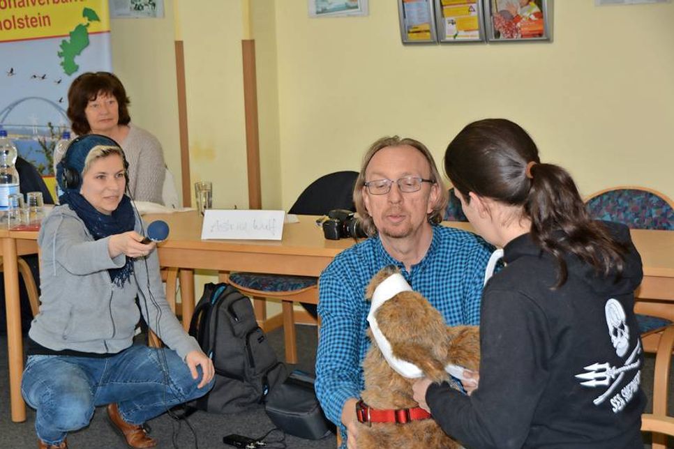 NDR-Redakteurin Astrid Wulf (links) fängt im Ton ein, wie Udo Galster und Ann-Christin Meyer (rechts)  einen Verband an der Schnauze von Übungshund ASBY anlegen, interessiert beobachtet von Ulrike Föse.