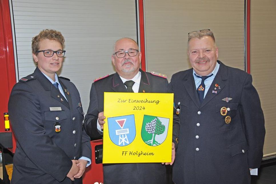 Michael Wilkat (m.) freut sich über das Geschenk der Partnerfeuerwehr aus Holzheim (Rheinland-Pfalz), das deren Wehrführer Dirk Weber und Stellvertreterin Sarah Kolter mitgebracht hatten: „…wird einen ehrenvollen Platz erhalten“.