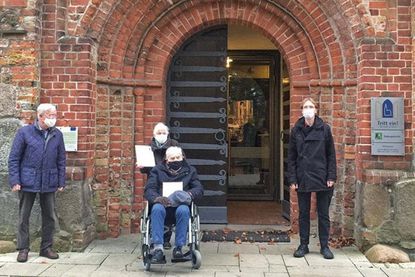 Hütedienstleiter Klaus Kasper (links) und Pastor Andreas Gruben (rechts) überreichen vor dem Kirchenportal der St.-Johannis-Kirche die Dankurkunden an Karin und Hans Peters.