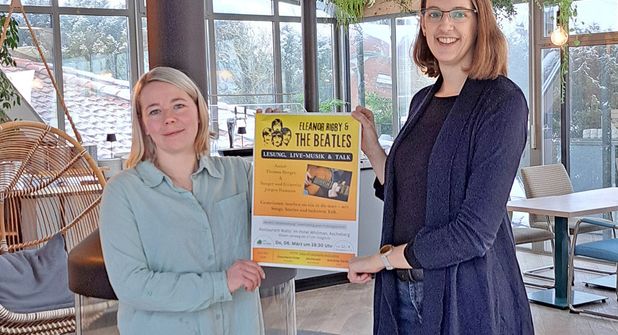 Sara Tesch (Restaurant Walts‘) und Alexa Gruthoff (Spiel und Freizeit für Generationen e.V.) präsentieren das Veranstaltungsplakat für den Beatlesabend am 6. März.